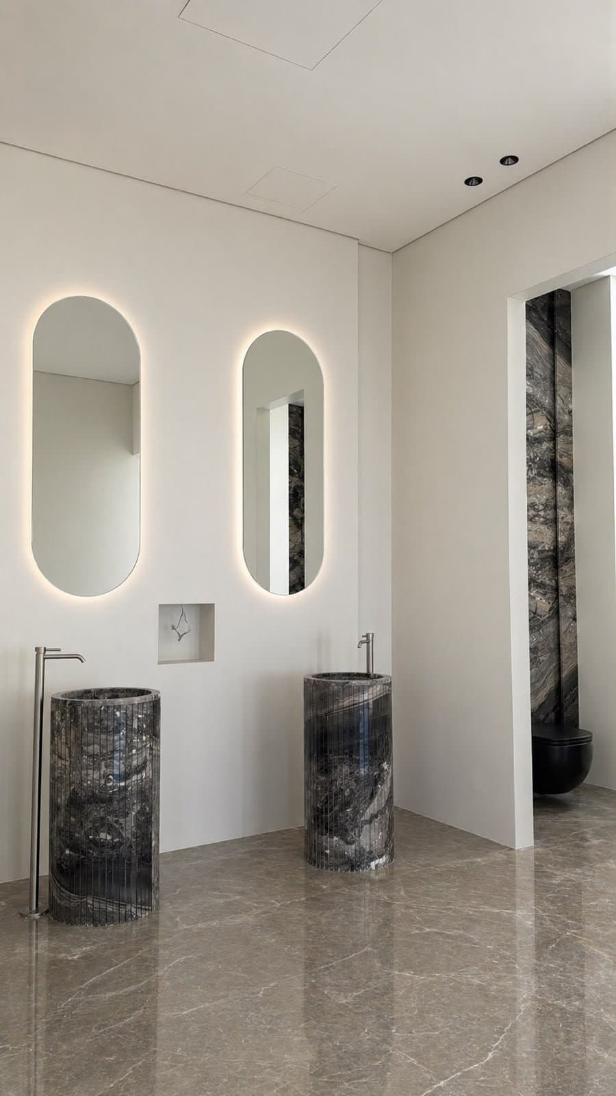 Dark stone cylindrical pedestal sinks beneath oval backlit mirrors